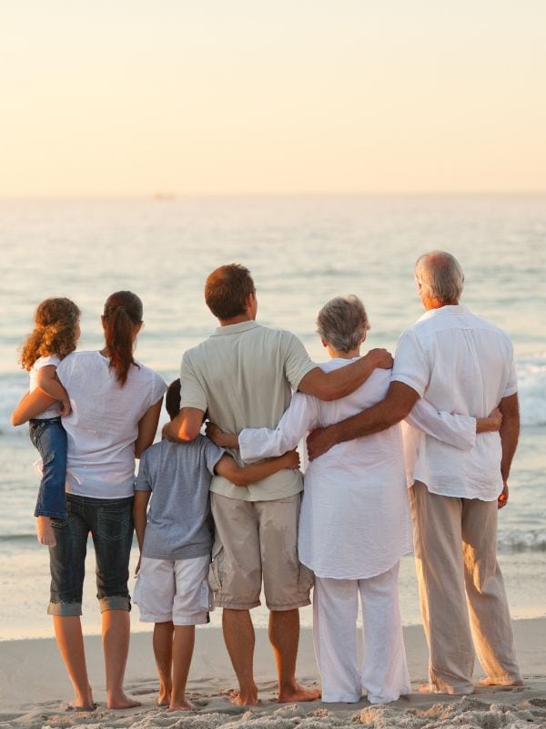 A multigenerational family stands on the beach at sunset, facing the ocean. The group includes grandparents, parents, and two young children. They are dressed in light-colored, casual clothing, with arms wrapped around each other, symbolizing closeness and togetherness. The calm sea and warm light create a peaceful, loving atmosphere.
