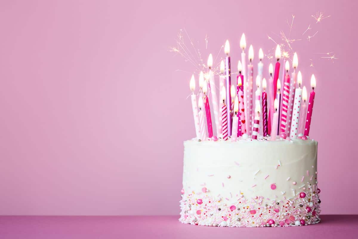 White frosted birthday cake decorated with pink and white sprinkles and topped with tall pink candles, some sparkling, set against a soft pink background.